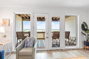 Quarto com sofá bege, mesa de madeira e vista para a praia através de grandes portas deslizantes.