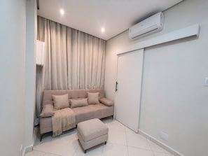 Una sala de estar moderna con un sofá color beige, un reposapiés a juego, un ventanal con cortinas y un equipo de aire acondicionado montado en la pared.
