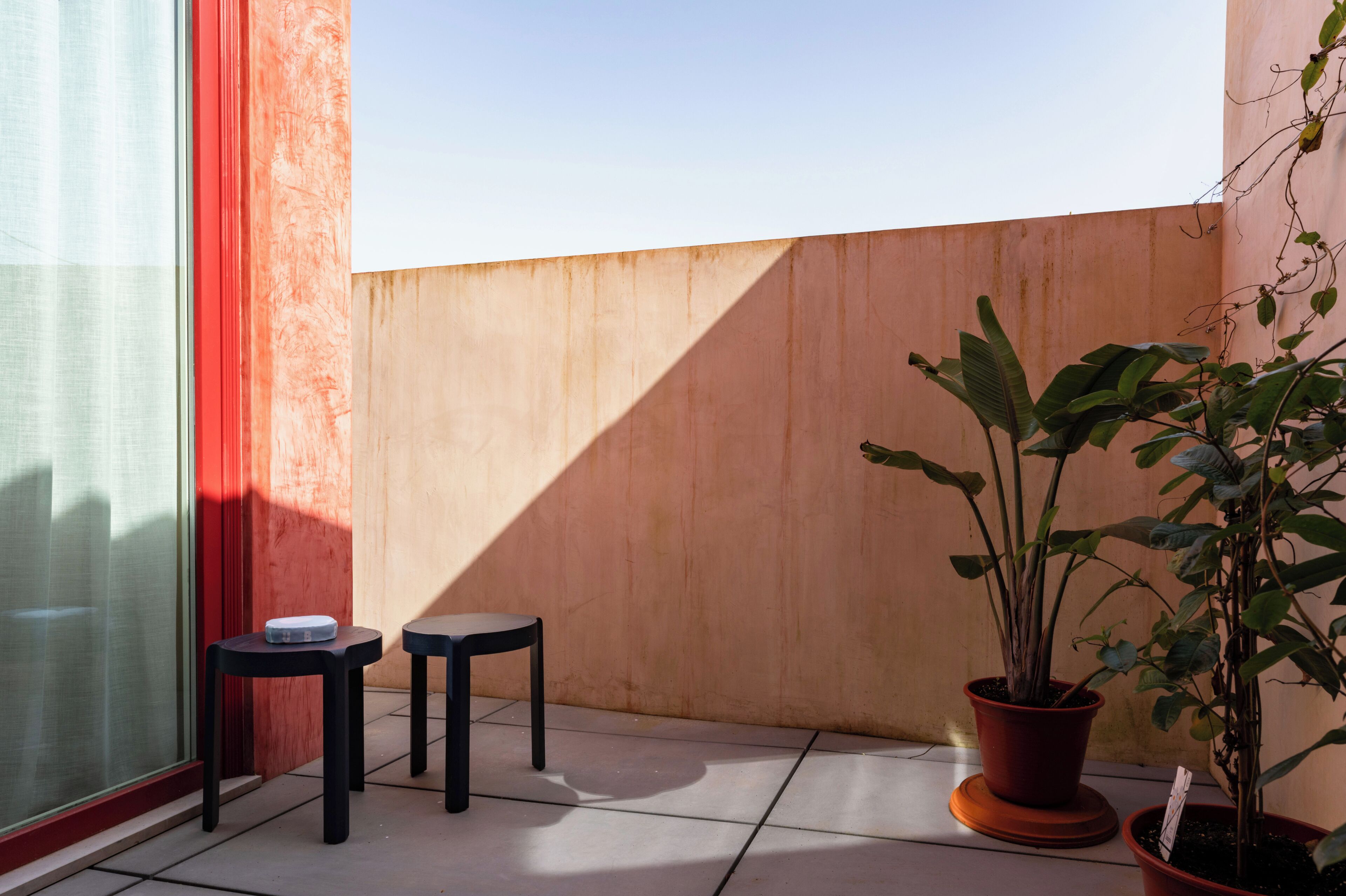 A balcony with two black stools, a potted plant, and a window with red curtains.