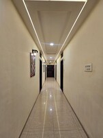 Un largo pasillo bien iluminado con suelo de baldosas, puertas a ambos lados y un cartel en la pared derecha.