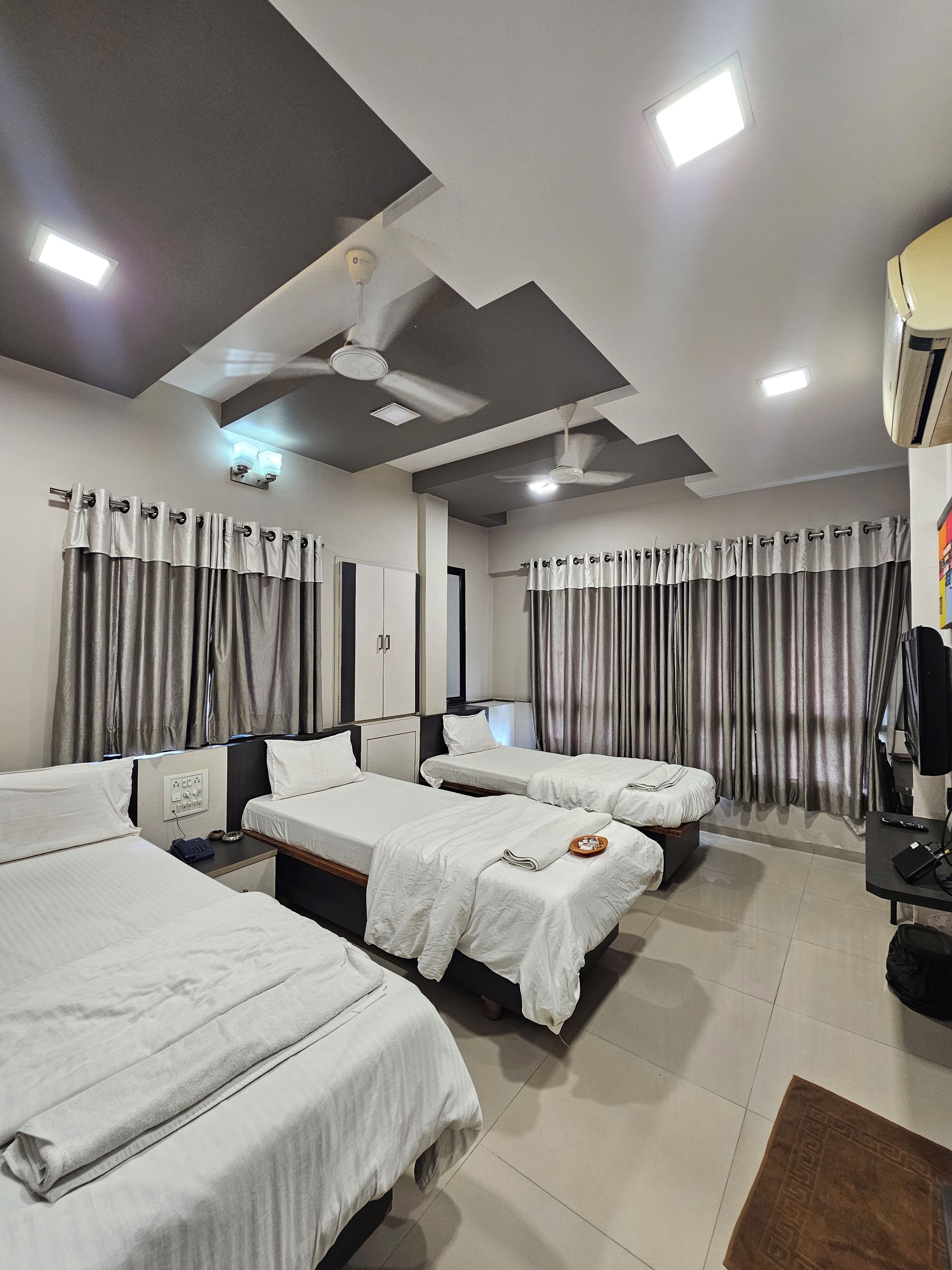 A hotel room with four single beds, a ceiling fan, recessed lighting, and a large window with curtains.