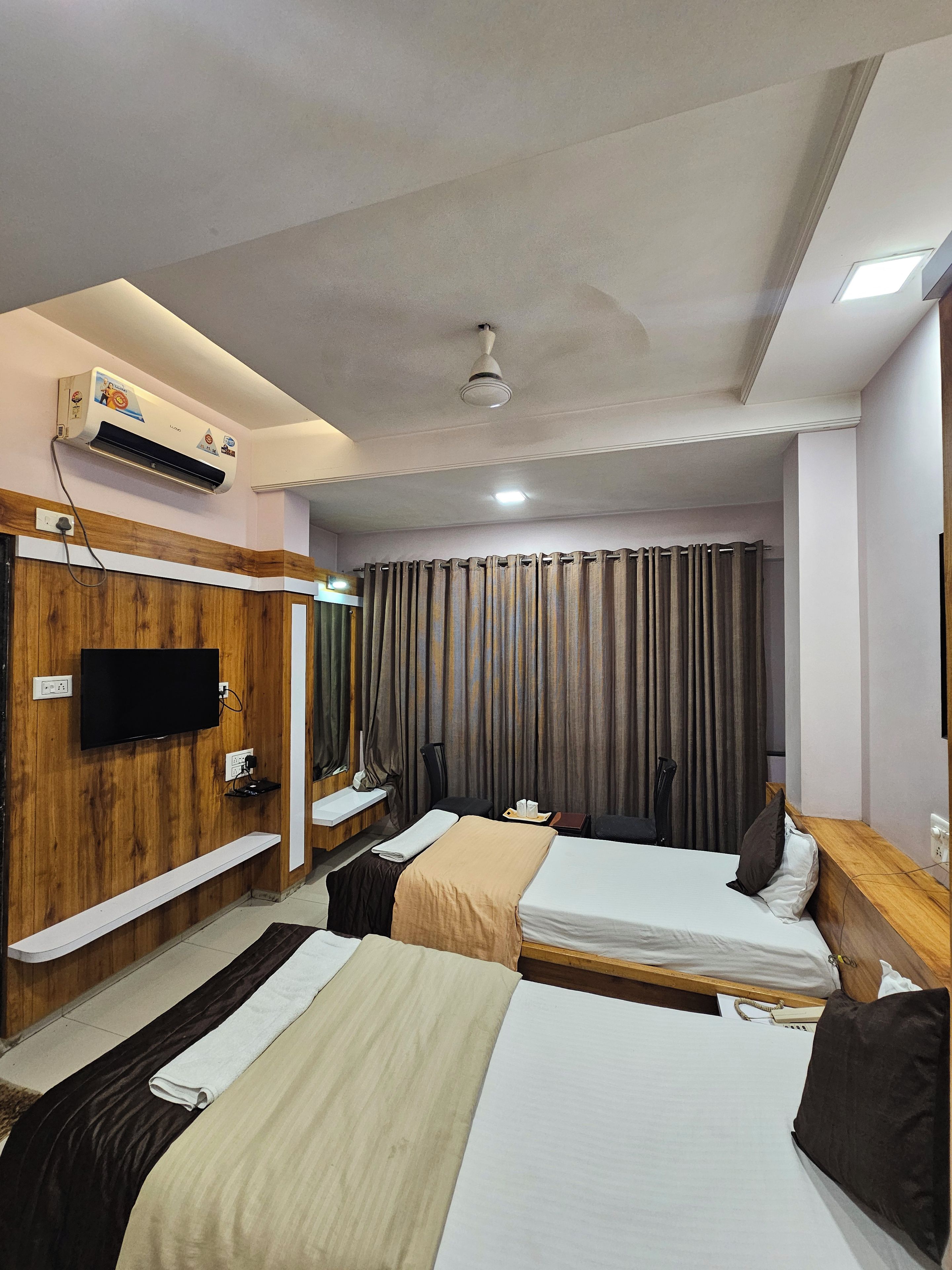 A hotel room with two beds, a TV, an air conditioning unit, a ceiling fan, and a large window with curtains.