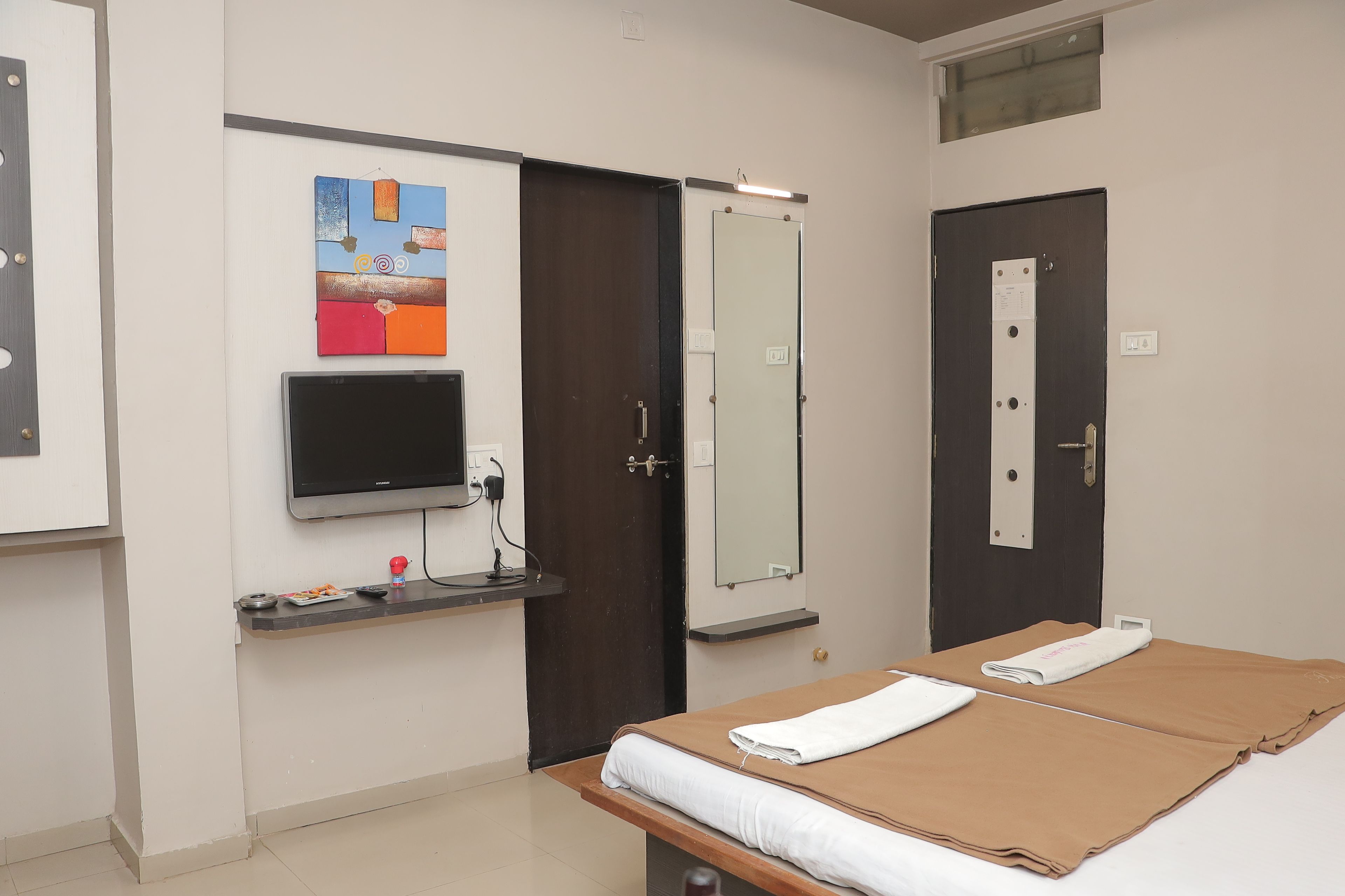 Une chambre avec un lit, un téléviseur fixé au mur, une porte avec un miroir et une petite étagère sur laquelle se trouve une télécommande.
