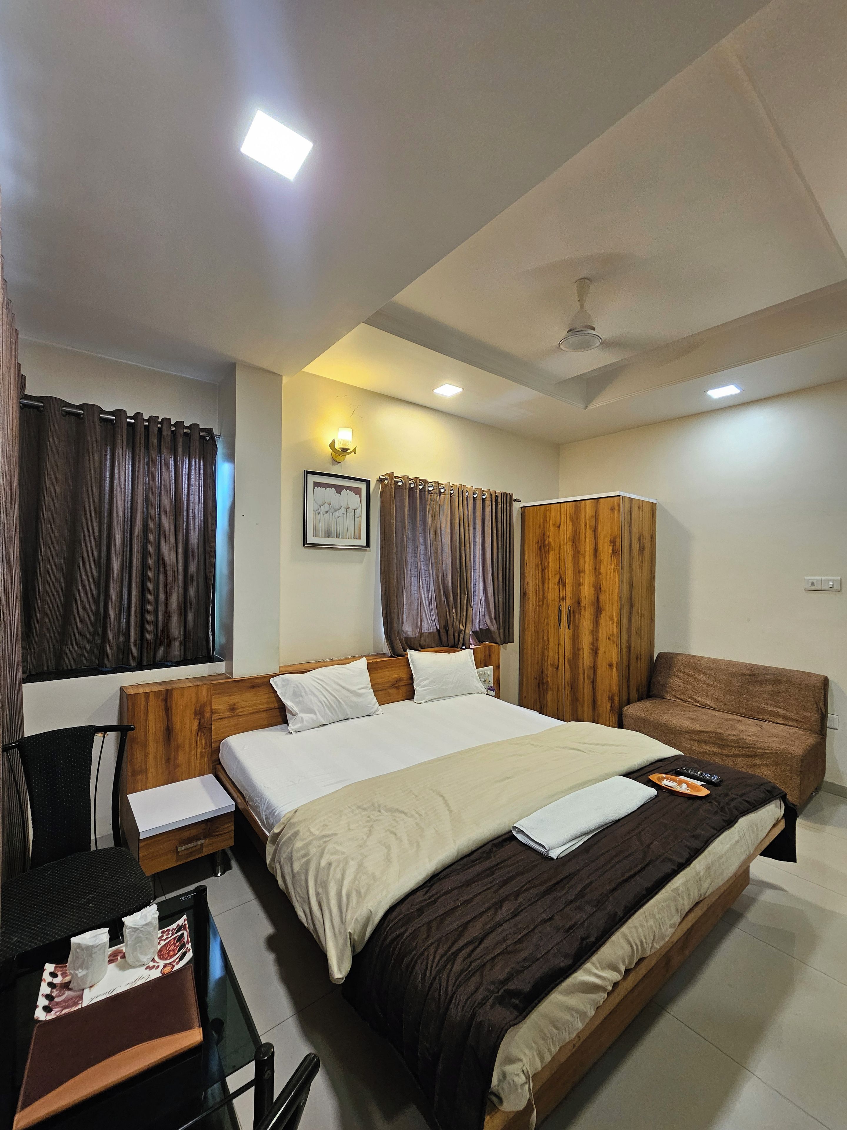 A hotel room with a large bed, a wooden wardrobe, a desk with a chair, and a small table with a tissue box and remote control.