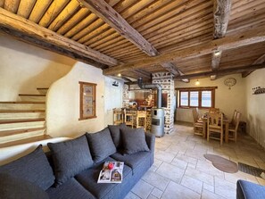 Ein gemütliches Interieur mit Holzbalkendecke, einer Couch mit Kissen, einem Essbereich mit Tisch und Stühlen sowie einem Steinkamin.