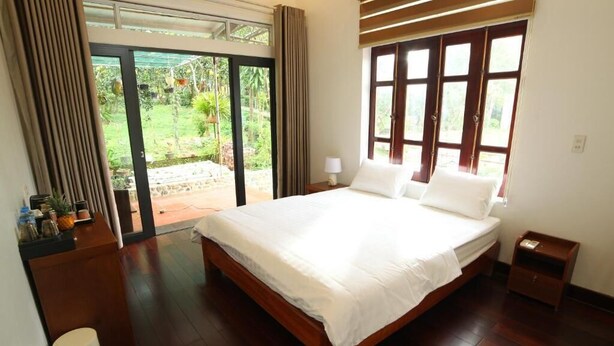 A bedroom with a large bed, a nightstand, a lamp, and a view of a garden through a sliding door. - Double Room with Garden View (Hà Nội)
