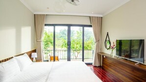 Ein Hotelzimmer mit einem großen Bett, einem Fernseher, einem Balkon mit Blick ins Grüne und einem Deckenventilator.