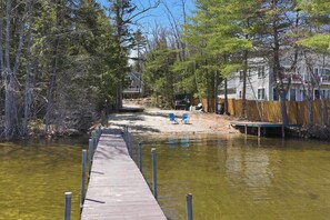Eine Holzstege führt zu einer Sandfläche mit Stühlen, umgeben von Bäumen und Häusern.