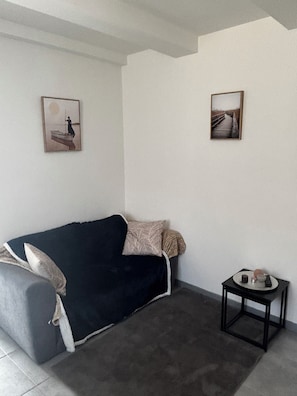 A room with a dark-colored sofa, a small side table with a remote and a decorative item, and two framed pictures on the wall.