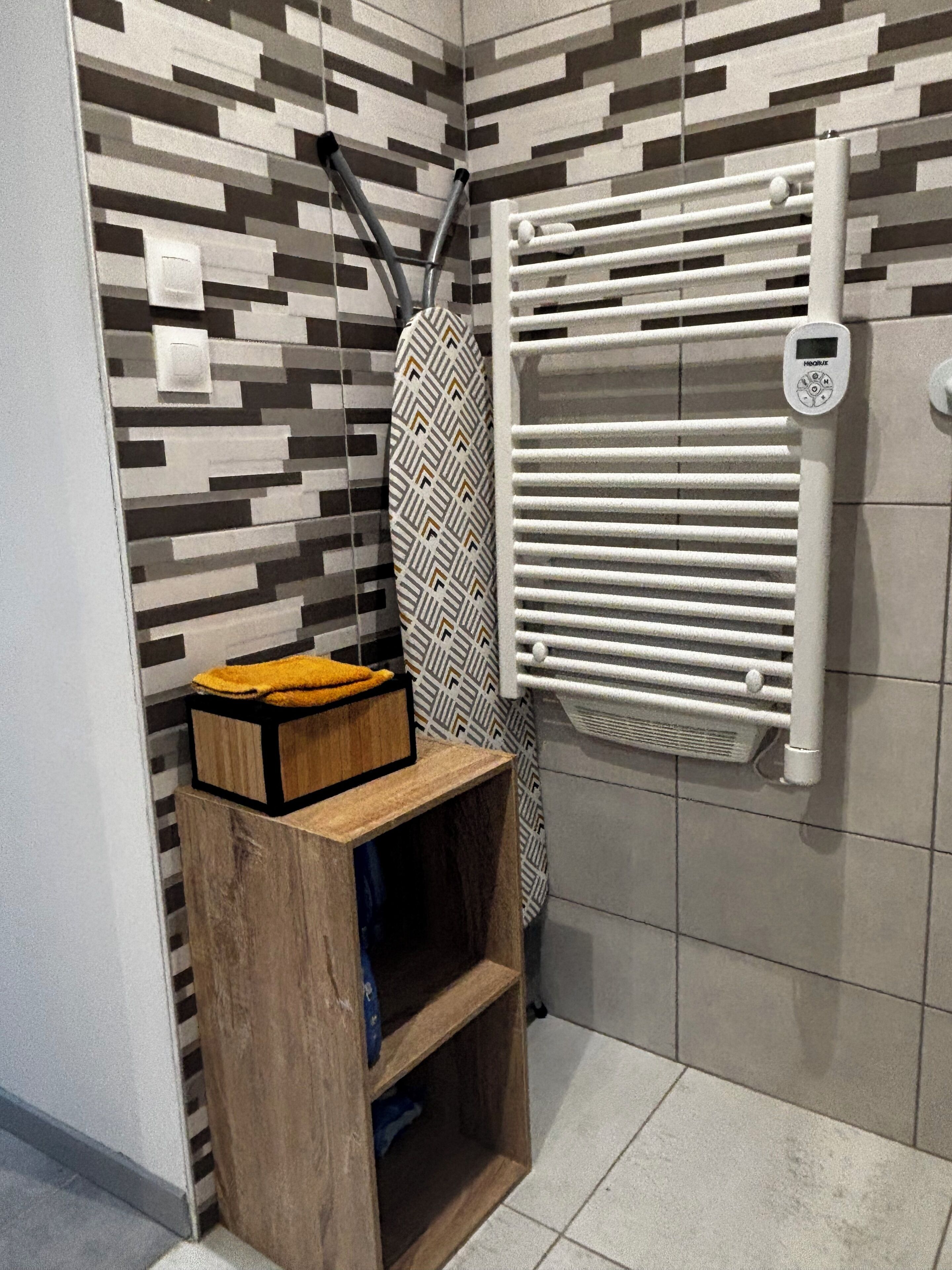 A bathroom with a patterned wall, a towel rack, a radiator, and a wooden shelf with a yellow towel.