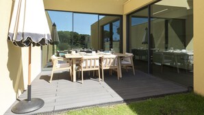 Un área de comedor al aire libre con una mesa y sillas de madera, una partición de vidrio y una sombrilla.