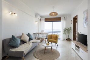A living room with a sofa, armchair, TV stand, and a dining area with a table and chairs. - Oceanides - Sea front with pool by HD (Quarteira)
