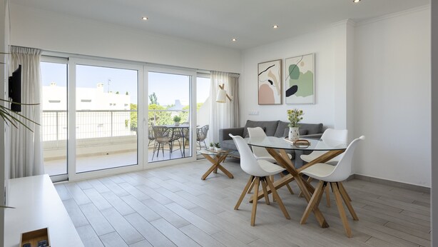 Un salon moderne avec une table à manger en verre, des chaises blanches, un canapé gris et un balcon aménagé avec du mobilier d’extérieur.