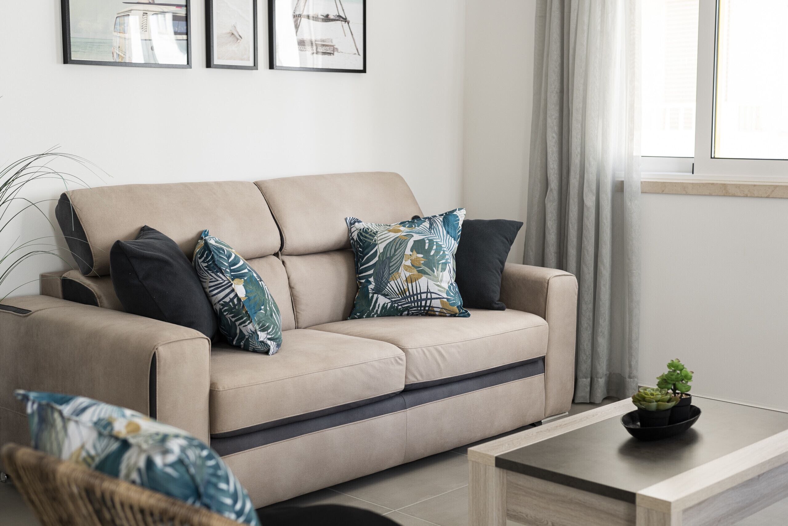 A beige sofa with patterned cushions, a small table with a plant, and framed artwork on the wall.