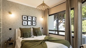 Een slaapkamer met een bed, een nachtkastje, een wandlamp, een raam met gordijnen, en ingelijste foto's aan de muur.