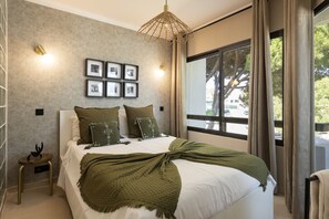A bedroom with a bed, a side table, a wall-mounted light, a window with curtains, and framed pictures on the wall. - Dona Marieta House by HD (Vilamoura)