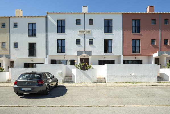 Fila de casas urbanas con un cartel que dice 'MOURALIZ' sobre la puerta central, un coche estacionado al frente y una pared blanca con una jardinera.