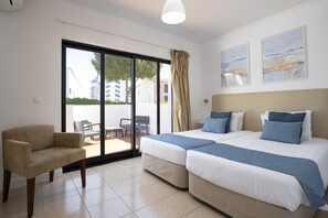 Habitación de hotel con dos camas, un sofá, un ventanal corredizo al exterior y dos cuadros en la pared.
