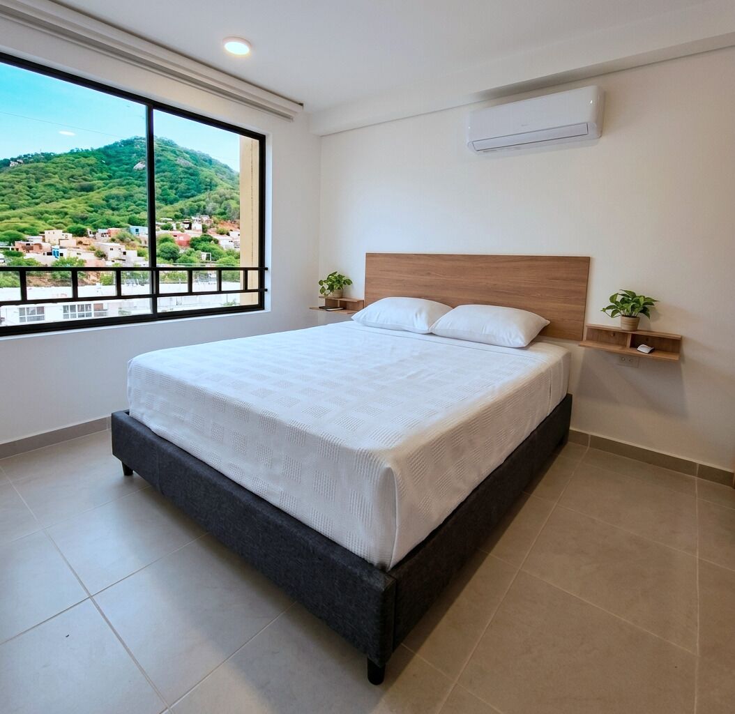 A modern bedroom with a large bed, a view of a green hillside, and a window with a black frame.