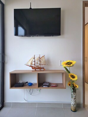 Ein modernes Wohnzimmer mit einem an der Wand montierten Flachbildfernseher, einem Holzregal mit elektronischen Geräten und einer Vase mit Sonnenblumen.