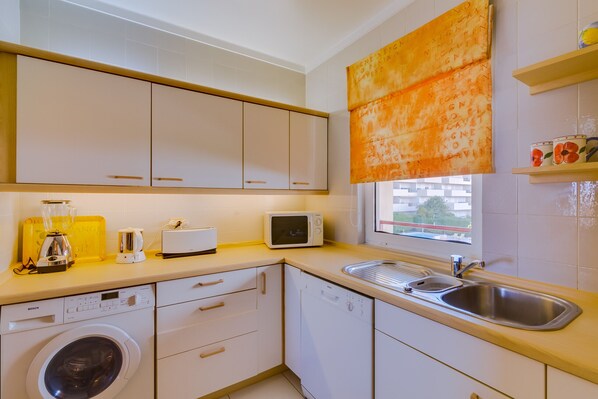 A compact kitchen with a washing machine, microwave, and a window with a view. - São Vicente - Vilamoura (Vilamoura)