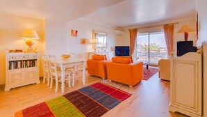 A living room with a TV, orange sofas, a white dining table, and a colorful rug. - São Vicente - Vilamoura (Vilamoura)