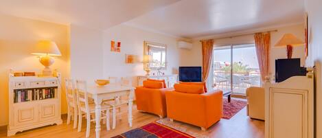 A living room with a TV, orange sofas, a white dining table, and a colorful rug.