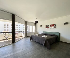 Ein modernes Hotelzimmer mit einem großen Bett, Blick auf die Stadt und einem Balkon mit Sitzgelegenheiten.