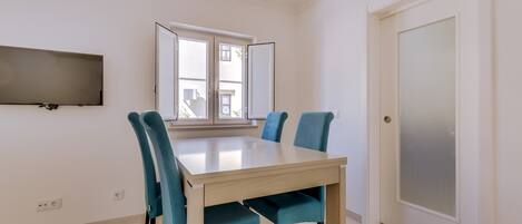 A dining area with a table and four chairs, a flat-screen TV on the wall, a window with a view, and a door with frosted glass.