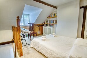 Ein Schlafzimmer mit schrägen Wänden, einem Bett mit weißen Leinen, einem Holzbett, einem Schreibtisch mit Tastatur und einem Bücherregal.