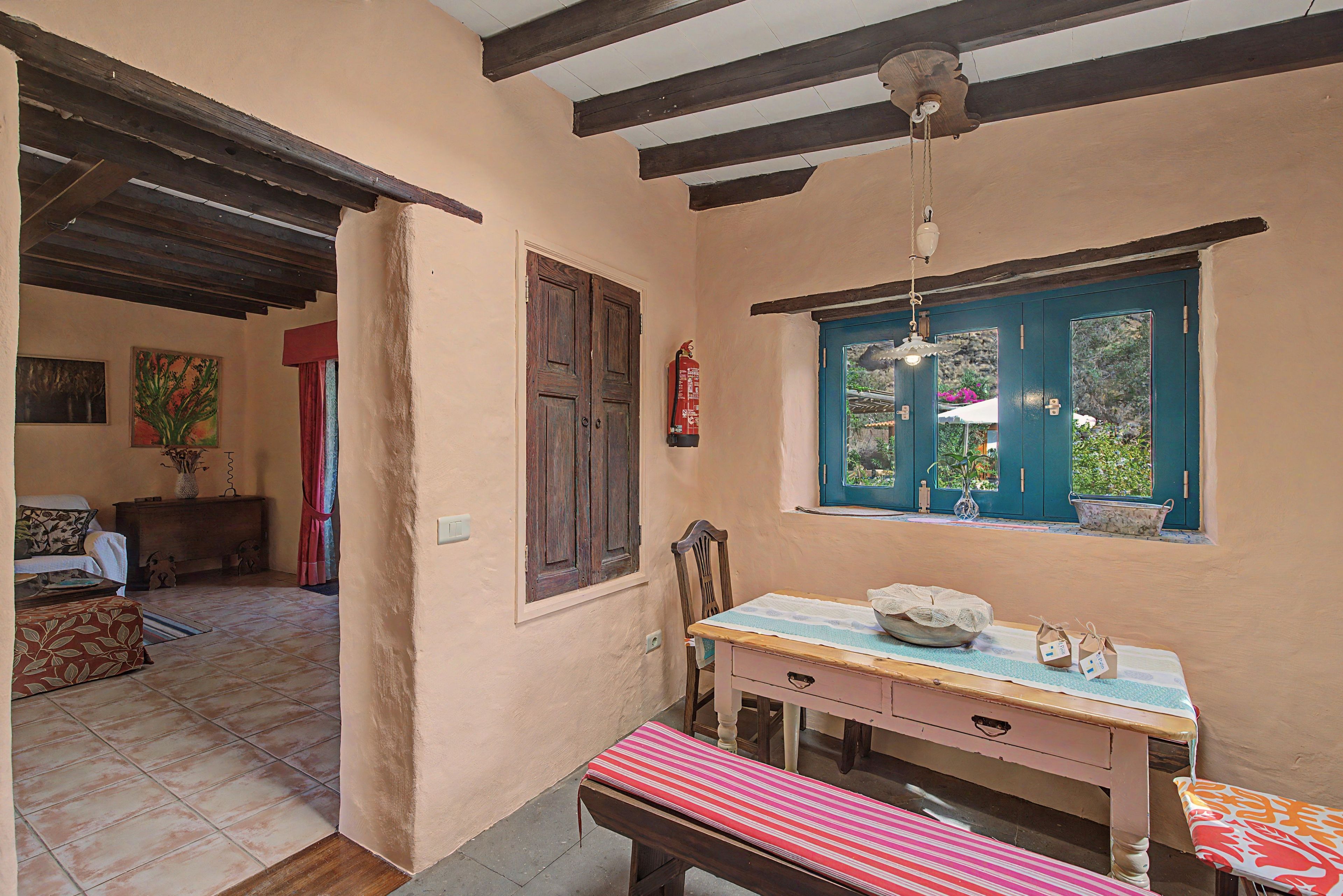 A room with a wooden ceiling, a table with a bowl and a vase, a window with a view of greenery, and a door with a red curtain.