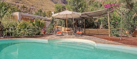 Ein Schwimmbecken mit einer Pergola und Stühlen, umgeben von üppiger Vegetation und einer Bergkulisse.