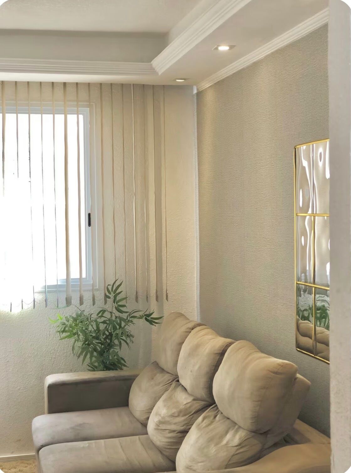 Una habitación con un sofá color beige, una planta en maceta, una ventana con persianas verticales y una obra de arte colgada en la pared.