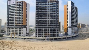 A modern high-rise building with multiple floors, a curved facade, and a surrounding sandy area. - New Studio w/ Balcony & Pool | Riviera Beachfront (Dubai)