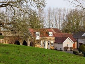 A stone building with a red-tiled roof, arched passageways, and a grassy foreground. - The Bothy (Knaresborough)