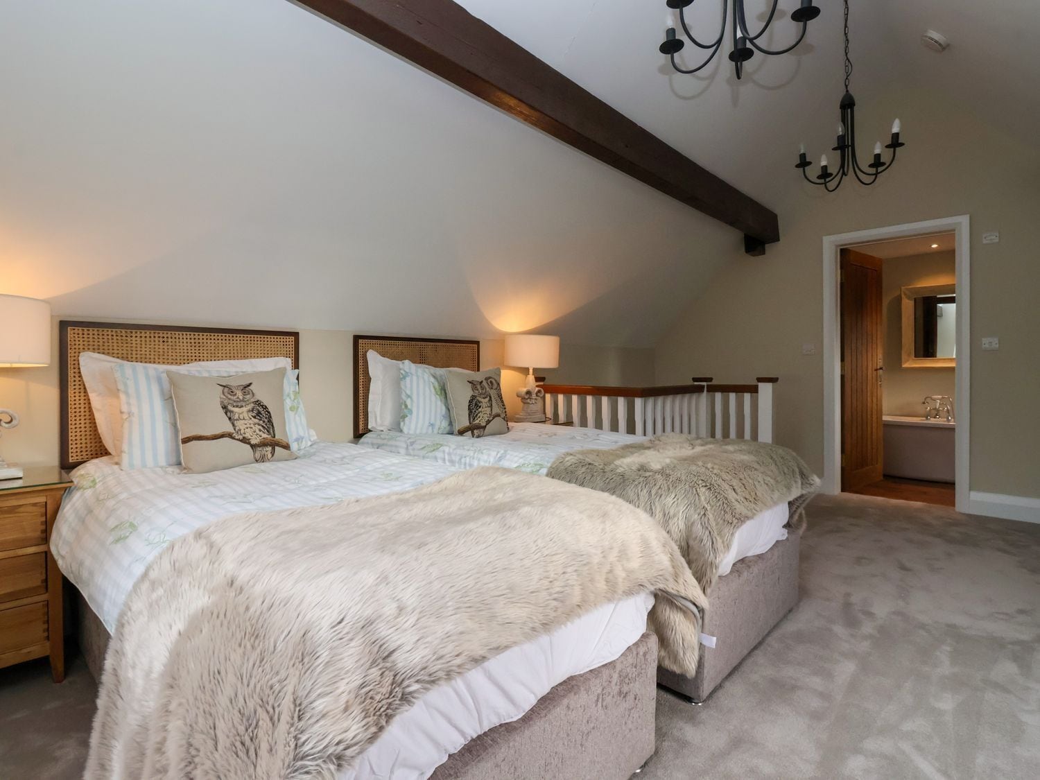 A bedroom with two beds, a chandelier, a wooden ceiling beam, and a bathroom visible through an open door.