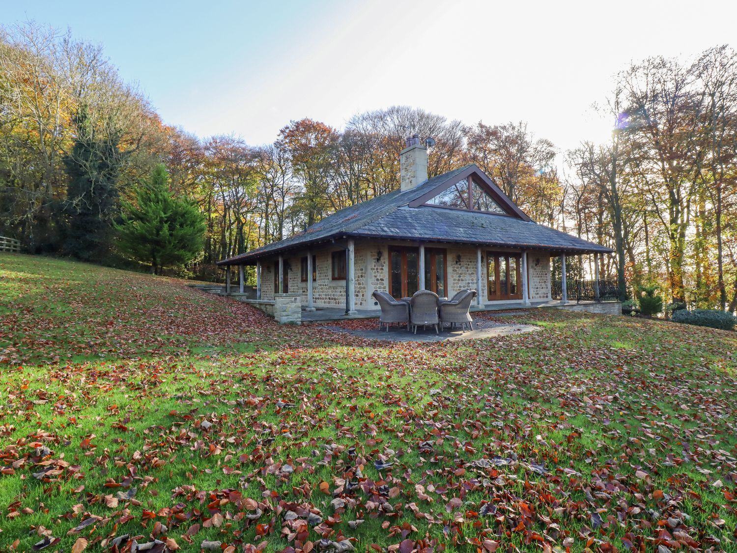 A stone building with a pitched roof, a covered patio with chairs, and a lawn with scattered leaves.