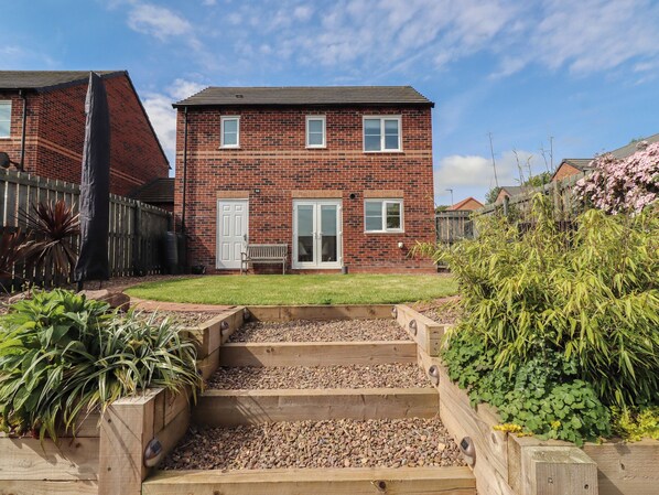 A two-story brick house with a front door, two windows, and a small front yard with a wooden bench and plants. - 26 Field Court (Wooler)