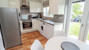 A modern kitchen with a refrigerator, oven, dishwasher, and washing machine. There is a dining table with chairs and a view of the outdoors through a glass door. - 26 Field Court (Wooler)