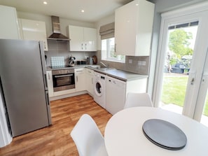 A modern kitchen with a refrigerator, oven, dishwasher, and washing machine. There is a dining table with chairs and a view of the outdoors through a glass door. - 26 Field Court (Wooler)