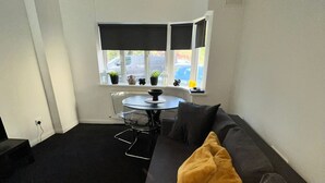 Una sala de estar moderna con alfombra negra, un sofá gris con un cojín amarillo, una mesa de vidrio y una ventana con persianas.