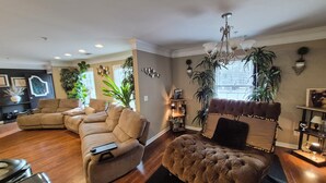 A living room with a sectional sofa, a tufted ottoman, a fireplace, and various plants. - The LaPearla Experience - A Hidden Gem (Riverdale)