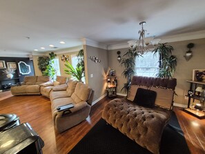 A living room with a sectional sofa, a tufted ottoman, a fireplace, and various plants.