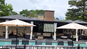 An outdoor pool area with lounge chairs and umbrellas, a brick building with a chimney, and a stone wall. - The LaPearla Experience - A Hidden Gem (Riverdale)