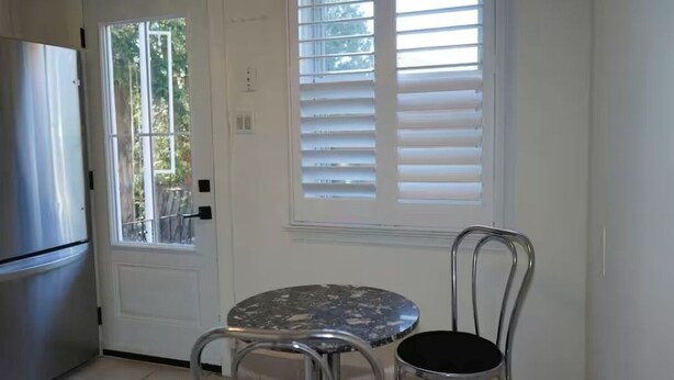 Una cocina con un refrigerador de acero inoxidable, una mesa redonda con una silla metálica y una ventana con persianas.