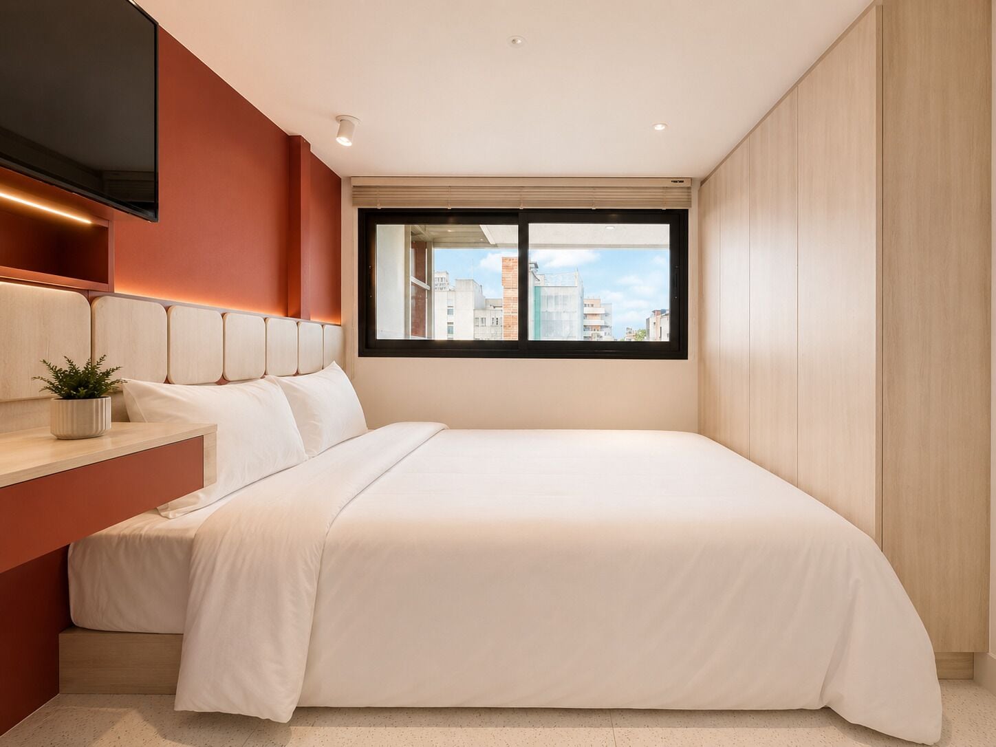 Une chambre d’hôtel moderne équipée d’un lit, d’une table de chevet avec une plante, d’une fenêtre donnant sur la ville et d’un téléviseur fixé au mur.