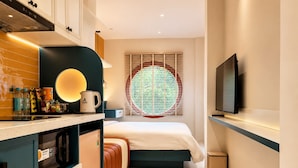 Quarto de hotel moderno com cozinha compacta, cama, televisão e janela com persianas.