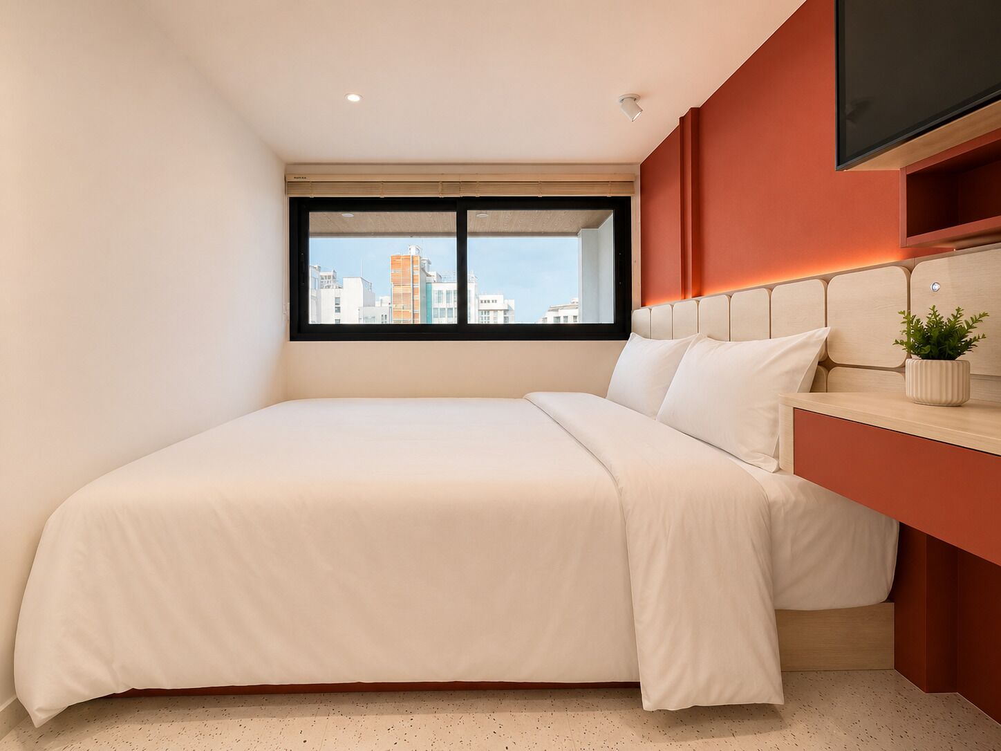 A modern hotel room with a large bed, a window with a city view, a mounted TV, and a small plant on the bedside table.