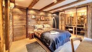 A bedroom with a large bed, wooden walls, a small side table, and a bathroom with a mirror and shelves. - Chalet, Ground floor, balcony, fireplace or stove, tv, ski locker, 380m², Saint Martin de Belleville (Saint-Martin-de-Belleville)