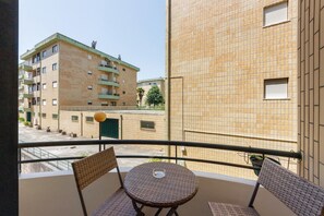 Un balcon avec des fauteuils en osier et une petite table, donnant sur une cour intérieure bordée d’immeubles et agrémentée d’un arbre verdoyant.